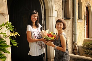 Zwei Frauen halten gemeinsam einen geflochtenen Weidenkorb mit Lebensmitteln und lächeln in die Kamera
