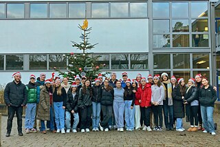 Schülerinnen und Schüler stehen vor einem weißen Gebäude mit vielen Fenstern. Sie tragen Weihnachtsmützen. Im Hintergrund steht ein festlich geschmückter Christbaum