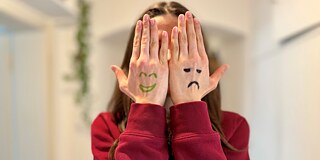 Eine junge Frau verdeckt ihr Gesicht mit den Händen. Auf den Handrücken sind links ein lächelndes Gesicht, rechts ein trauriges Gesicht gemalt.