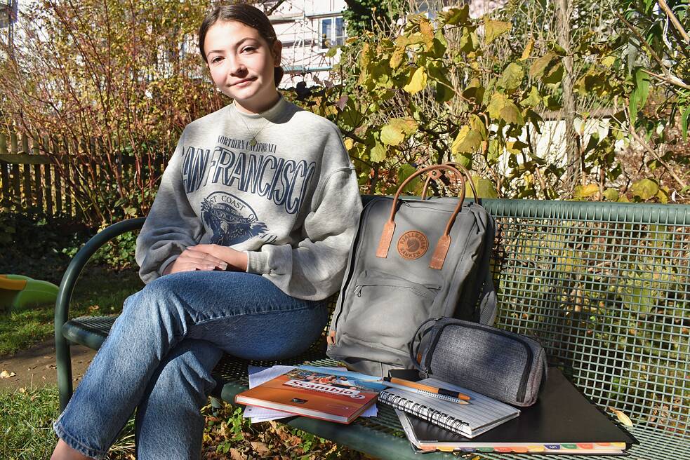 Marie-Sophie sitzt mit ihrem Schulrucksack und dem Inhalt ihres Rucksacks auf einer Bank und lächelt in die Kamera.
