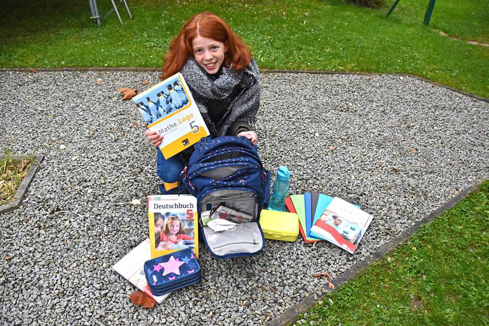 Mädchen sitzt mit ihrer Schultasche auf dem Boden und hält ihr Mathebuch in die Luft