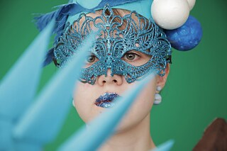 Nahaufnahme einer Person mit hellblauer Maske, blauem Kopfschmuck und blau geschminkten Lippen