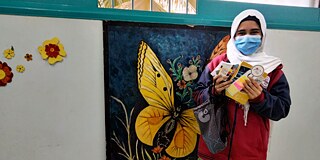 Schülerin mit Maske im Schulgebäude hält Büromaterialien und steht vor einem Plakat mit Schmetterlingen
