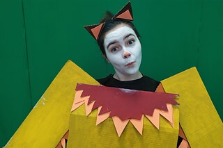 Nahaufnahme des Oberkörpers einer Person in einem gelben Kostüm mit rotem Kragen und weiß geschminktem Gesicht vor einem Greenscreen