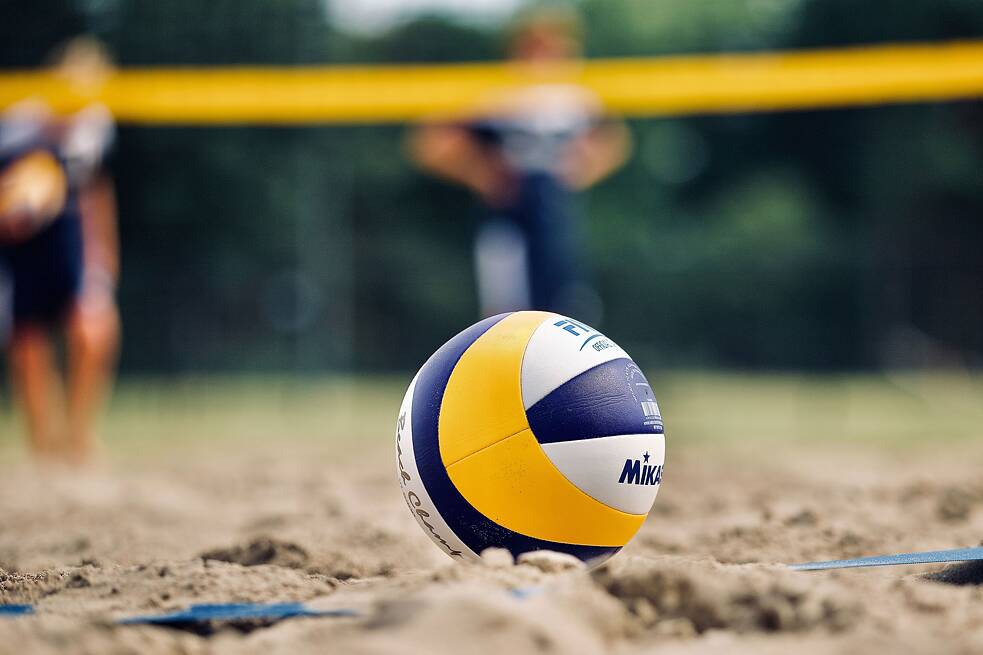 Im Vordergrund ein Volleyball im Sand, im Hintergrund mehrere Personen und ein Volleyballnetz