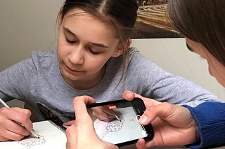 Schülerin malt, während eine Frau vor ihr sitzt und sie aufnimmt