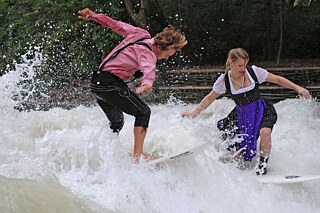 Zwei Personen in bayerischen Trachten surfen auf der Eisbachwelle in München