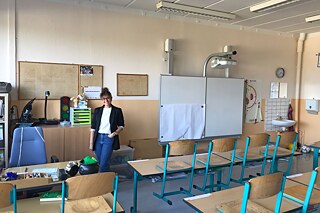 Heidi steht in einem leeren Klassenzimmer mit hochgestellten Stühlen am Pult