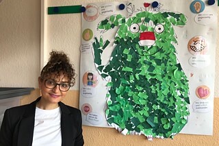 Heidi steht vor einem Plakat, auf dem ein gebasteltes grünes Monster befestigt ist und lächelt