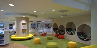 Bunte Bibliothek, an der der Seite eine Wand mit Kreisen, in denen man sitzen kann. Einige Schulkinder sitzen dort und lesen.
