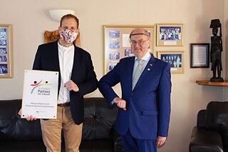 Zwei Männer mit Mund-Nasen-Schutz berühren sich mit den Ellenbogen. Der linke Mann hält die PASCH-Plakette