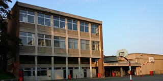 Gelbes Gebäude, im Vordergrund ein Basketballplatz mit Basketballkorb