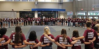 Schülerinnen und Schüler in einheitlichen dunkelroten T-Shirts mit der Aufschrift „Decide Commit Succeed.“ stehen im Kreis im Schulgebäude