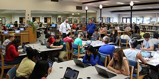 Schülerinnen und Schüler sitzen mit Lehrkraft an Laptops in der Bibliothek und lernen