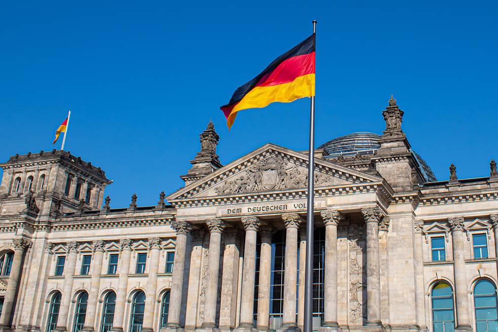 Der deutsche Reichstag, eine Deutschladflagge im Vordergrund, blauer Himmel.