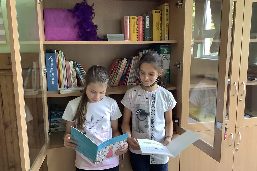 Zwei Mädchen halten jeweils ein Buch in den Händen und stehen vor einer Vitrine mit Büchern