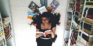 Frau liegt auf einem Stapel Bücher in einer Bibliothek auf dem Boden und liest ein Buch. 