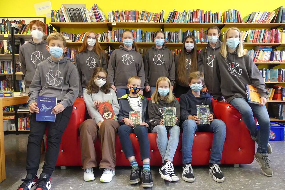 Gruppenbild von Schülerinnen und Schüler in den gleichen grauen Pullis vor einem großen Bücherregal