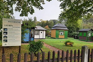 Bunt gestrichene Gartenlauben im Hintergrund, im Vordergrund ein Schild mit dem Titel "Historische Gartenlauben"