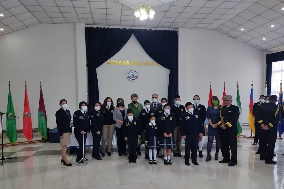 Gruppenfoto in einer Aula mit weißen Wänden, im Hintergrund Flagge von Bolivien