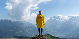 Frau in gelbem Regenmantel steht auf einem Felsen und schaut auf eine Landschaft