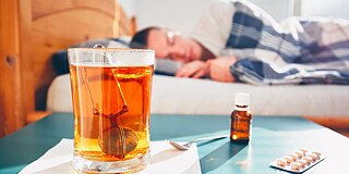 Eine männliche Person liegt im Bett, im Vordergrund ein Tee und ein Blister mit Tabletten