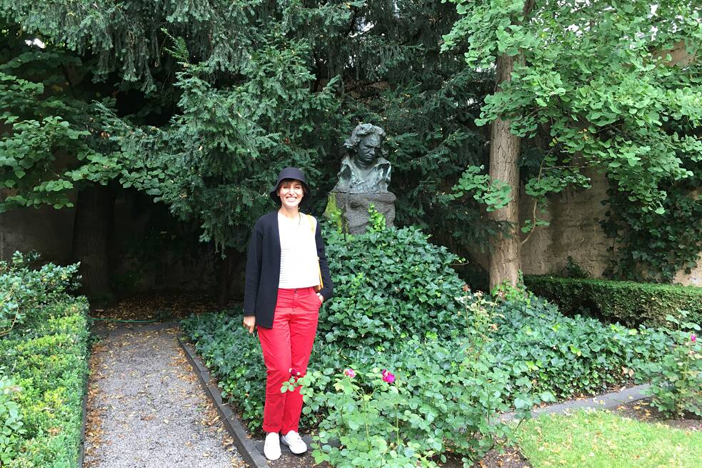 Eine Frau in einer roten Hose und einem weißen Oberteil steht in einem Park neben einer Büste