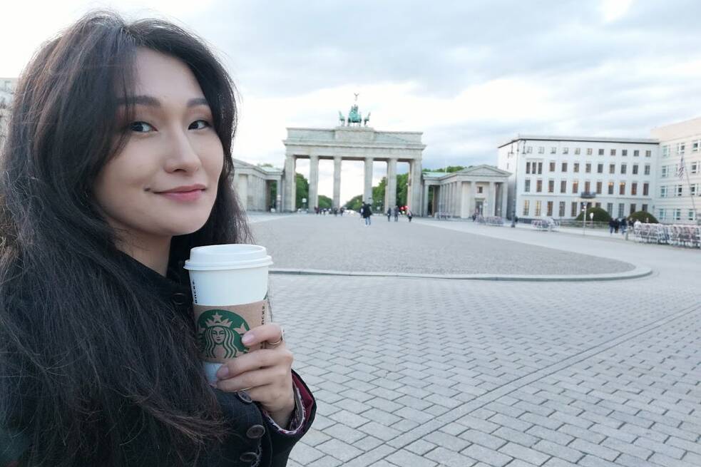 Eine Frau macht ein Selfie von sich mit einem To Go-Kaffee vor dem Brandenburger Tor