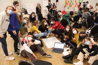 Schülerinnen und Schüler sitzen auf dem Boden in einem Klassenraum und arbeiten an Plakaten