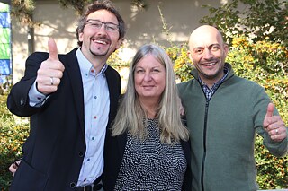 Drei Personen stehen für ein Foto zusammen, die beiden Männer links und rechts halten jeweils einen Daumen hoch