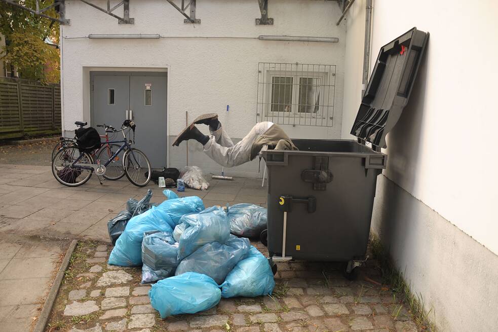 Mann hängt kopfüber in einer Mülltonne, davor ein paar blaue Müllsäcke