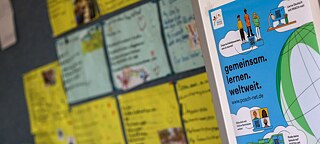 PASCH-net-Poster mit verschiedenen Grafiken (Weltkugel, Personen, Schulgebäude) an einer Pinnwand in einem Klassenzimmer