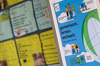 PASCH-net-Poster zum Download PASCH-net-Poster mit verschiedenen Grafiken (Weltkugel, Personen, Schulgebäude) an einer Pinnwand in einem Klassenzimmer