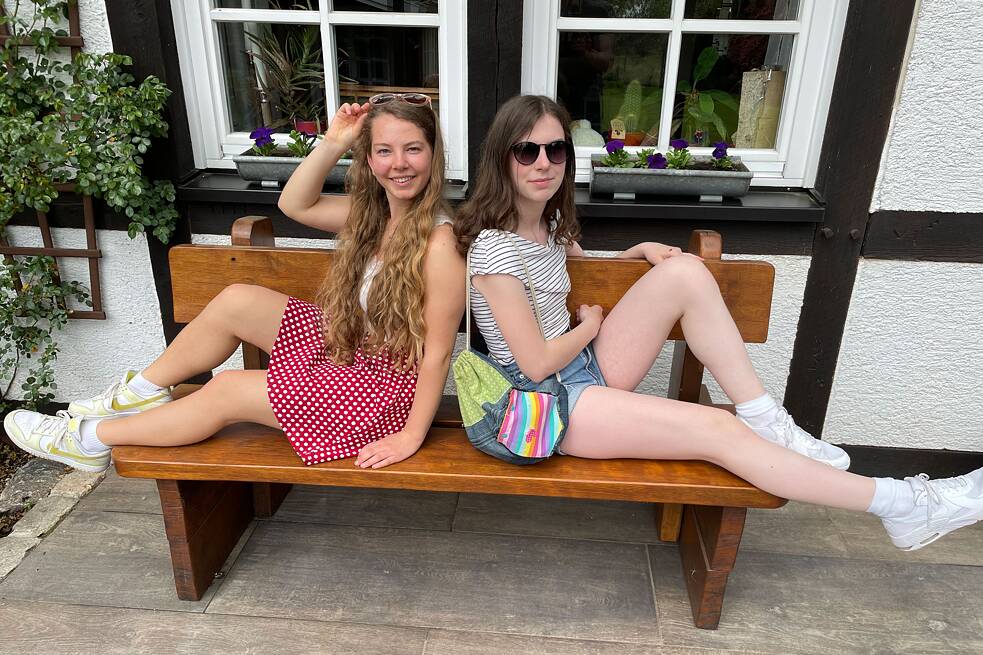 Zwei junge Frauen sitzen auf einer Bank vor einem Fenster mit dem Rücken zueinander