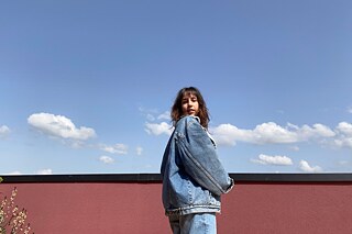 Frau in Jeansjacke und Jeanshose auf einem Balkon seitlich fotografiert
