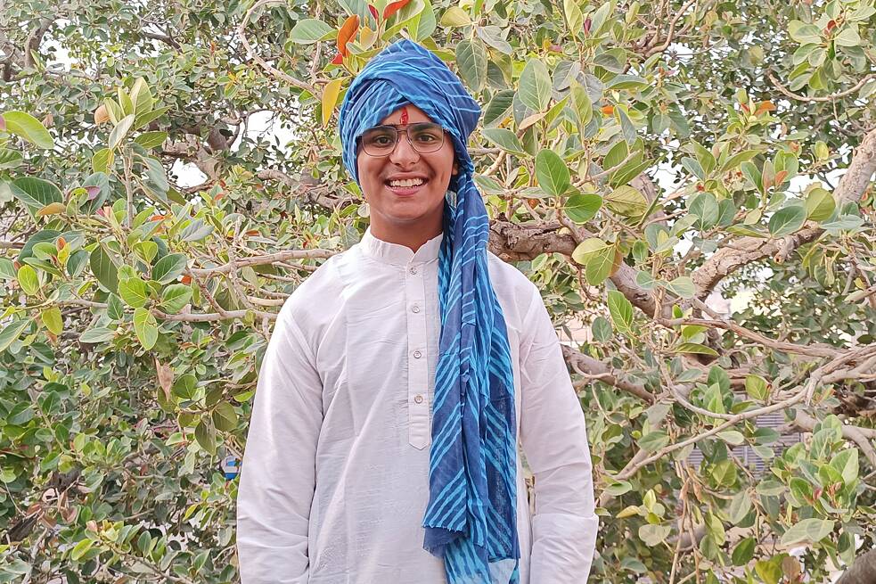 Junge in weißer Kurta mit Knöpfen und einem blauen Turban vor einer Hecke fotografiert