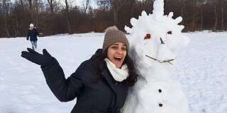 Frau mit Winterjacke, Handschuhen und Mütze posiert neben einem großen Schneemann