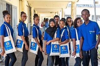 Mehrere Schülerinnen und Schüler in blauen Oberteilen und beigen Jutebeuteln stehen zusammen für ein Gruppenfoto