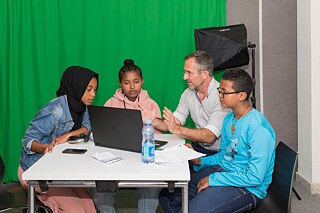 Drei Jugendliche sitzen mit einem  Mann an einem Tisch und schauen auf einen Laptop