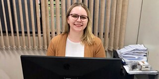 Pamela © privat Frau mit Brille, weißem T-Shirt und beiger Jacke sitzt vor einem Rechner im einem Büro und lächelt in die Kamera