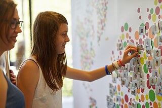 Die interaktive Weltkarte von PASCH-net. Zwei Personen pinnen ein Foto an eine Leinwand mit der PASCH-Weltkarte.