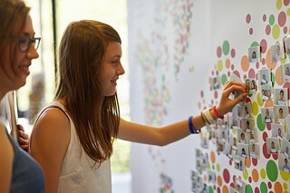 Zwei Personen pinnen ein Foto an eine Leinwand mit der PASCH-Weltkarte.