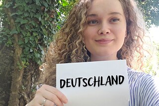#PASCHinWien auf der IDT 2022 Eine Hand hält ein Kärtchen mit dem Hashtag PASCH in Wien. Im Hintergrund ist ein leicht bewölkter Himmel.
