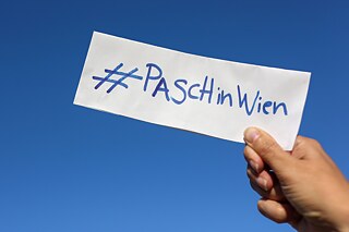 #PASCH in Wien auf der IDT Eine Hand hält ein Kärtchen mit dem Hashtag PASCH in Wien. Im Hintergrund ist ein wolkenloser Himmel.