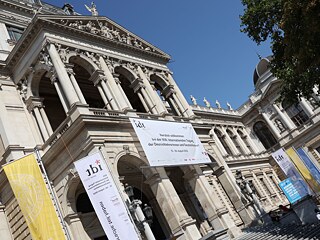 Der Haupteingang zur IDT: Außenansicht des Hauptgebäudes der Uni Wien, davor Banner und Flaggen der IDT und Uni Wien.