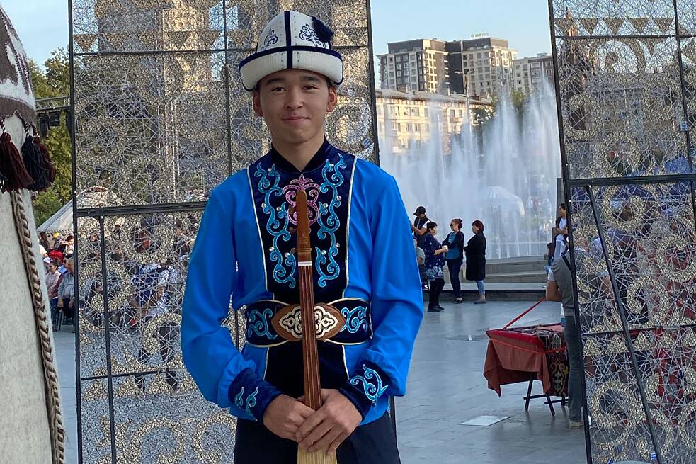 Junge in einem blauen, langärmeligen Hemd, weißem Hut und einem Zupfinstrument vor einem Springbrunnen fotografiert