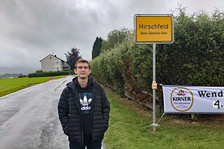 Ein junger Mann steht vor dem Ortsschild Hirschfeld, Rhein-Hunsrück-Kreis