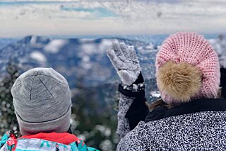 Zwei Personen mit Pommelmütze und dicken Jacken von hinten auf einem Berg