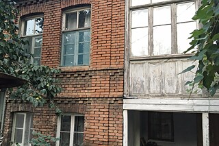 Rot-braunes, altes Backsteingebäude mit vier Fenstern. Daneben eine Holzfassade mit blickdichten Fenstern
