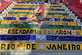 Frau in rotem Oberteil sitzt auf einer Treppe aus bunten Kacheln, welche in den Farben der brasilianischen Flagge dekoriert ist